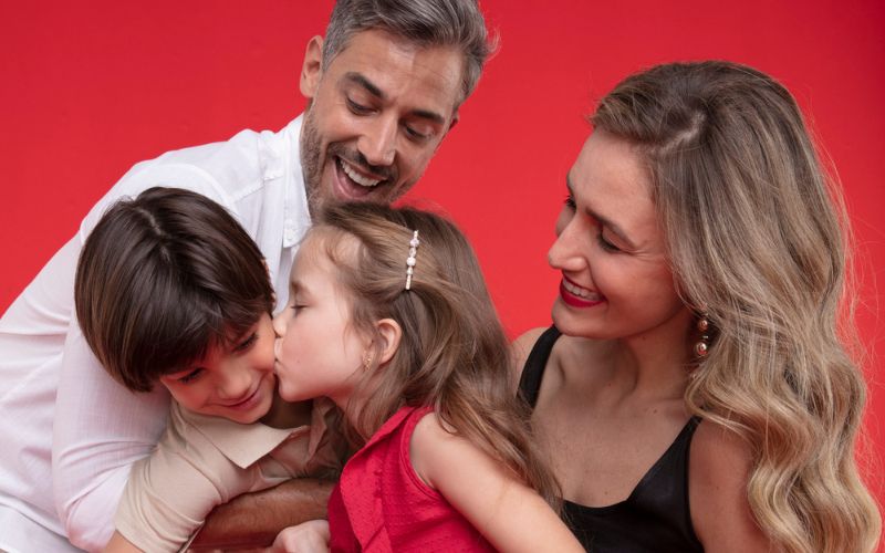 Familia tirando uma foto a menina esta dando um beijo no rosto do seu irmão