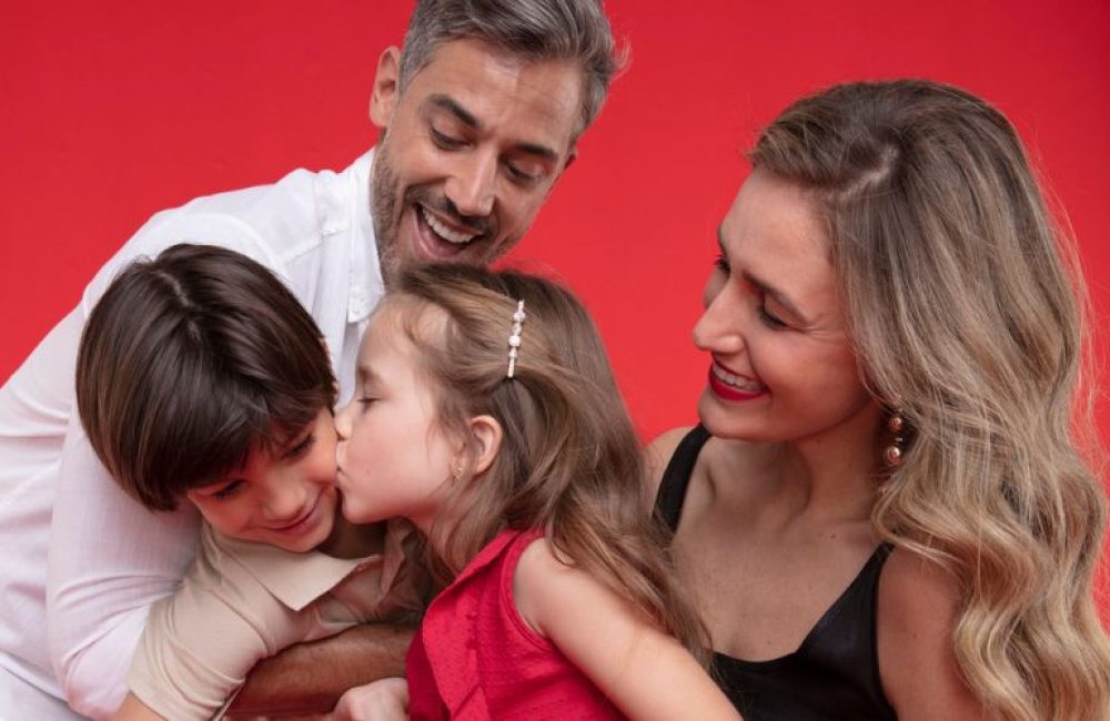 Familia tirando uma foto a menina esta dando um beijo no rosto do seu irmão